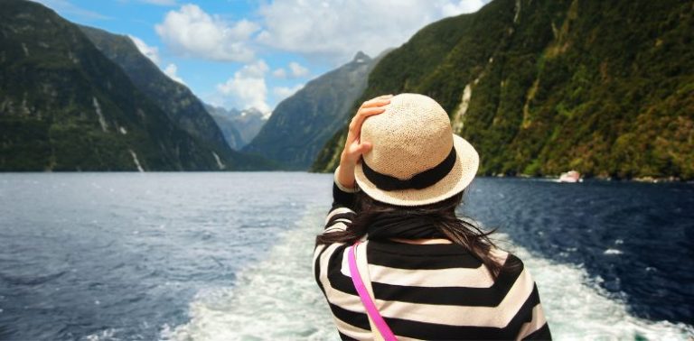 A view of the back of a woman, wearing a hat and long-sleeved shirt, looking off the side of a cruise ship at the water and mountains.
