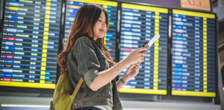 A woman looking at a plane ticket in front of the departures board at an airport.