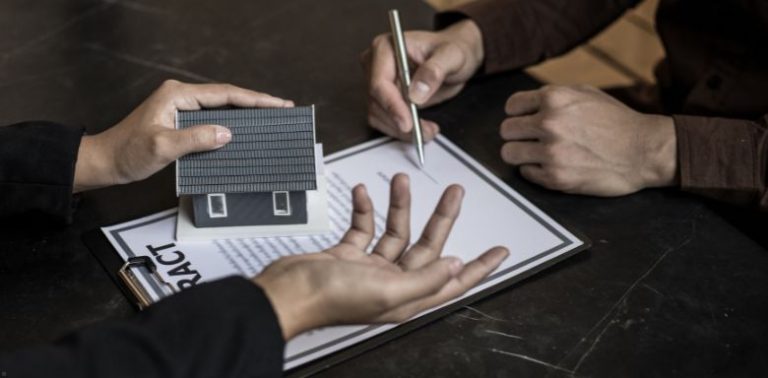 A person signing a contract. There is another set of hands holding a small model home.