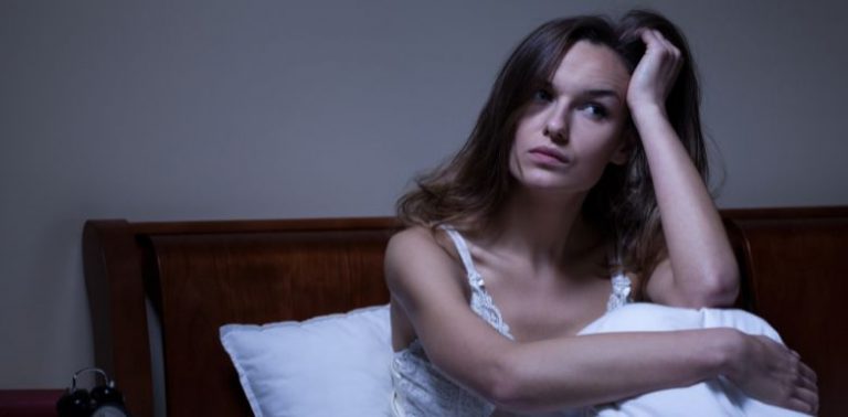 A woman sitting up in bed with a hand on her head. She is having a hard time sleeping.