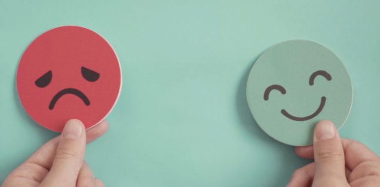 Fingers holding two paper cut-outs, one is a red sad face and the other is a teal happy face.