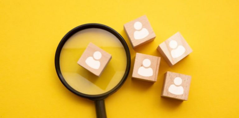 Small wooden squares with white people painted on them. One is under a magnifying glass. It has a bright yellow background.