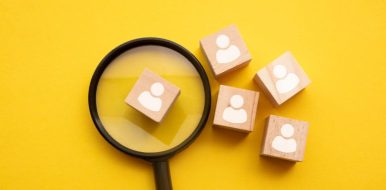 Small wooden squares with white people painted on them. One is under a magnifying glass. It has a bright yellow background.
