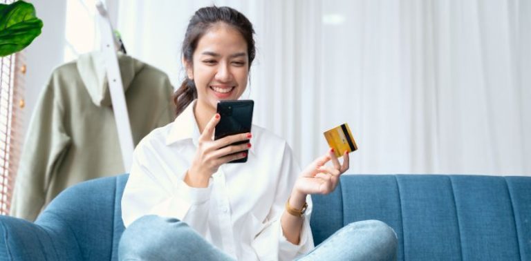A woman sitting on a couch holding her phone in one hand, and her credit card in another.
